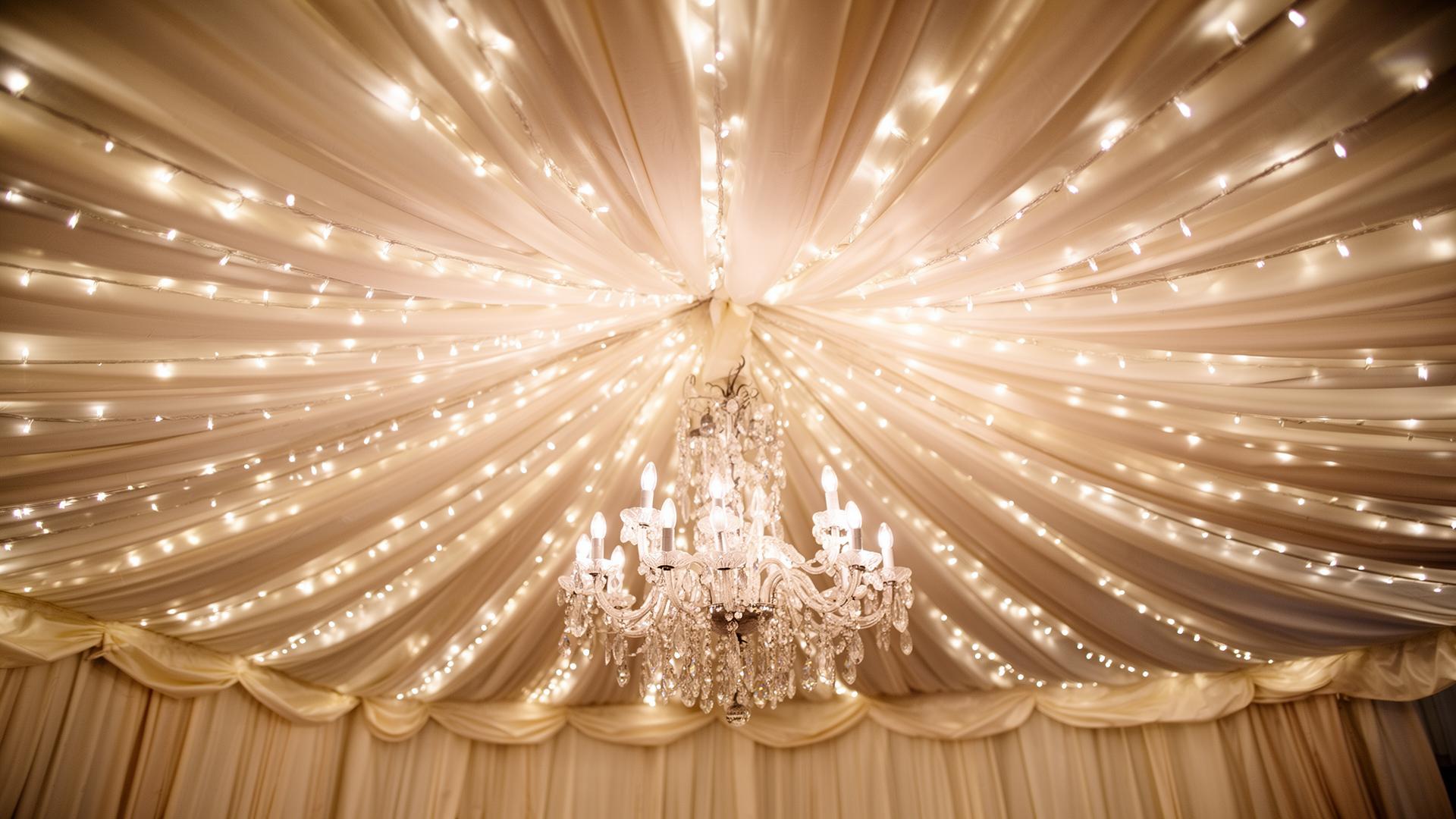 Detail of a Raphoe Marquee Hire interior showing pleated ivory silk ceiling linings, a starlight fairy-light canopy and a hanging crystal chandelier above a wedding reception